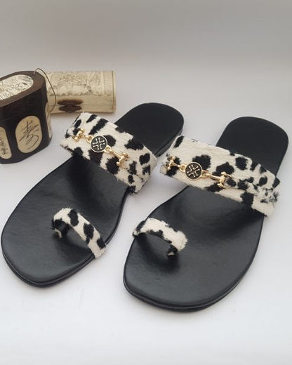 A pair of black velvet flip flops with white leopard print straps and decorative buckles, displayed alongside a wooden box possibly containing the packaging.
