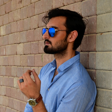 Man wearing sunglasses and a blue shirt leaning against a brick wall.