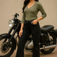 Woman in green top and black pants standing next to a motorcycle on a beige background