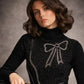 Woman wearing a black sweater with a bow design, holding a black handbag.