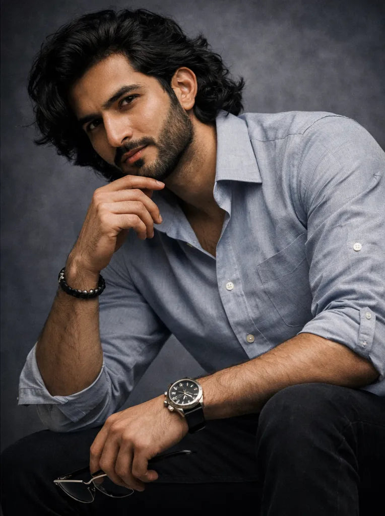 Man wearing a light blue shirt and black pants with a watch, sitting against a dark background.