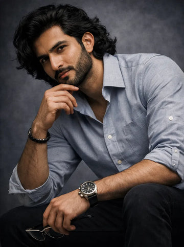 Man wearing a light blue shirt and black pants with a watch, sitting against a dark background.