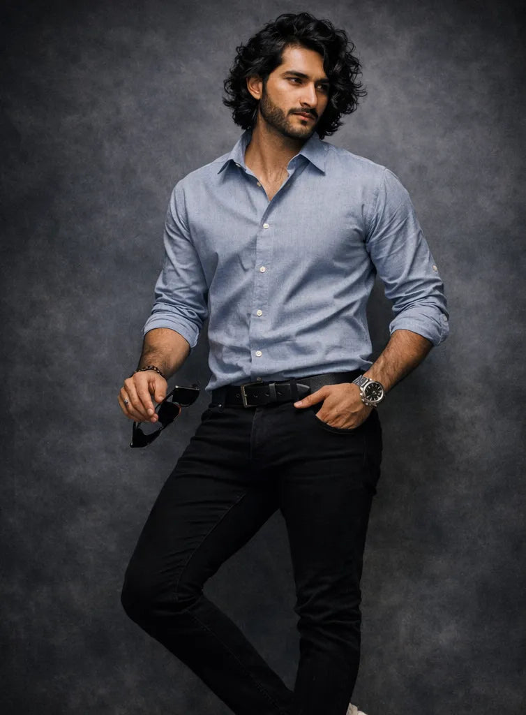 Man wearing a light blue shirt and black pants with a watch, sitting against a dark background.