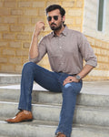 A man wearing a light brown Linen shirt, blue jeans, and brown leather dress shoes sits on concrete steps in front of a yellow brick wall.