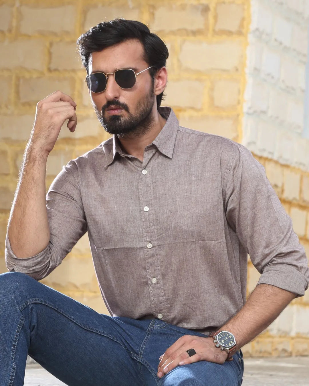 A man wearing a light brown linen shirt and blue jeans, sitting against a light-colored brick wall background.
