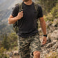 Man hiking in a mountainous area wearing a black t-shirt, camouflage shorts, and a green backpack.