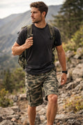 Man hiking in a mountainous area wearing a black t-shirt, camouflage shorts, and a green backpack.