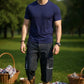 Man in navy blue t-shirt and dark shorts standing in a park with picnic baskets and blankets.