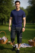 Man in navy blue t-shirt and dark shorts standing in a park with picnic baskets and blankets.