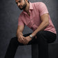 Man wearing a pink shirt and black pants sitting on a dark background