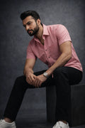 Man wearing a pink shirt and black pants sitting on a dark background