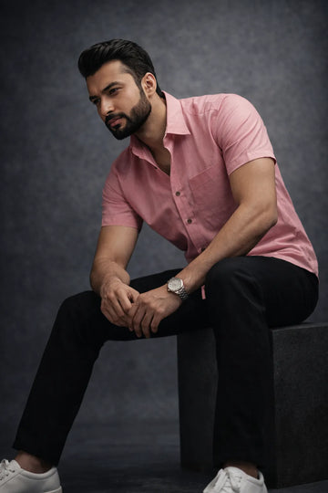 Man wearing a pink shirt and black pants sitting on a dark background