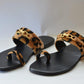 A pair of leopard printed velvet flip flops with a decorative buckle on a white background.