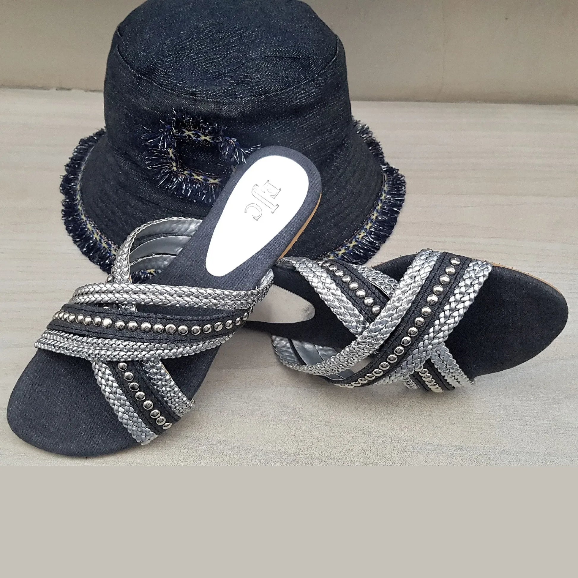Black sandals with silver embellishments and a matching black hat on a light wooden surface.