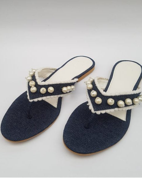 A pair of navy blue denim flip-flops embellished with white lace and pearls, displayed against a white background.