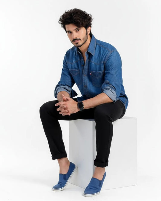 Man wearing a blue denim shirt and black pants sitting on a white cube against a white background