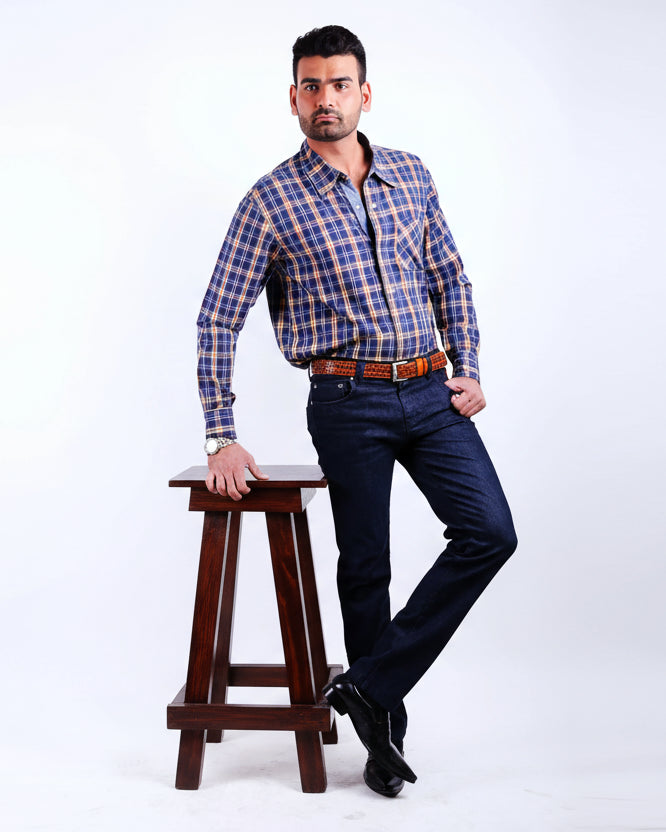 Man wearing a blue plaid shirt and dark jeans sitting on a wooden stool against a white background