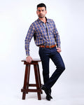 Man wearing a blue and red checkered shirt and dark jeans standing on a wooden stool against a white background