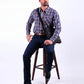 Man wearing a blue plaid shirt and dark jeans sitting on a wooden stool against a white background