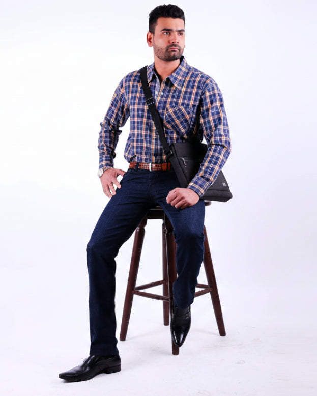 Man wearing a blue plaid shirt and dark jeans sitting on a wooden stool against a white background