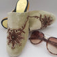 A pair of fawn velvet mule shoes with brown embroidery, displayed alongside a pair of sunglasses.
