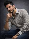 Man wearing a light gray shirt and dark jeans with a watch, sitting against a dark background