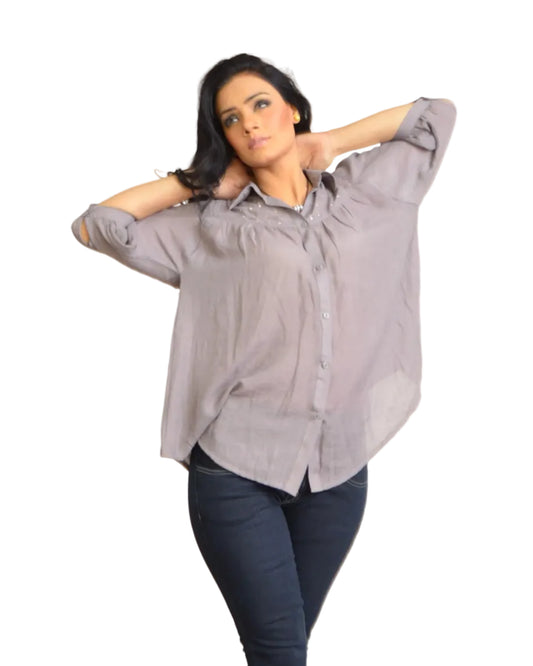 Woman wearing a gray button-up shirt and dark jeans standing against a plain wall.