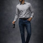 Man wearing a light gray shirt, dark jeans, and black shoes against a dark background