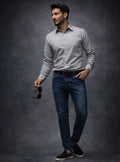 Man wearing a light gray shirt, dark jeans, and black shoes against a dark background