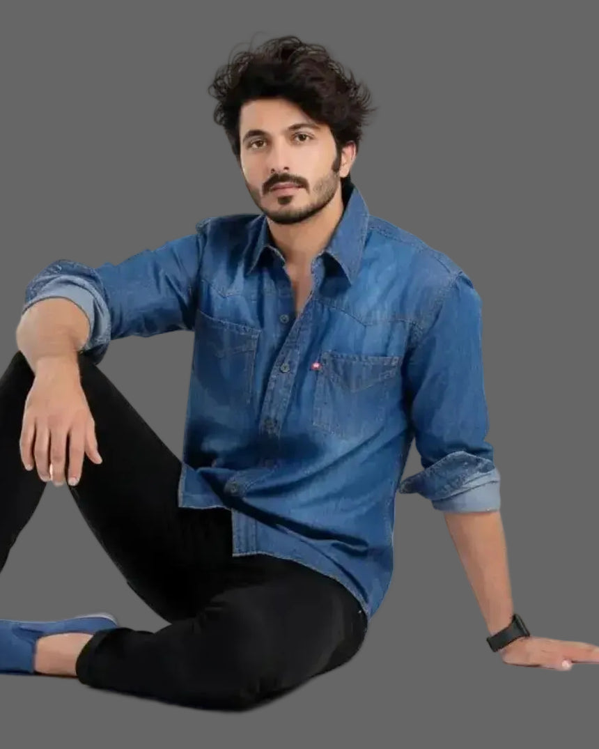 Man wearing a blue denim shirt and black pants sitting on a white background