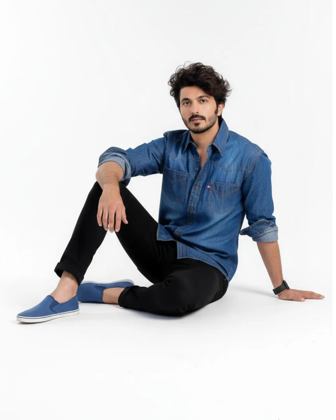 Man wearing a blue denim shirt and black pants sitting on a white background