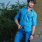 Man wearing a blue denim shirt and jeans standing outdoors with greenery and a wall in the background.