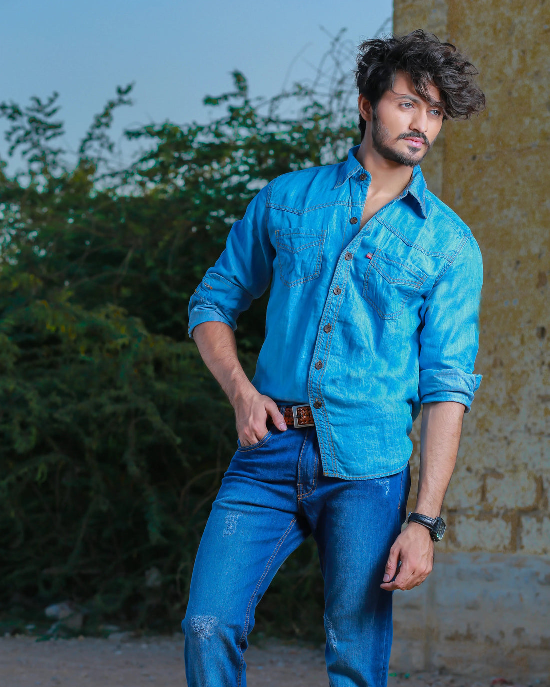 Man wearing a blue denim shirt and jeans standing outdoors with greenery and a wall in the background.