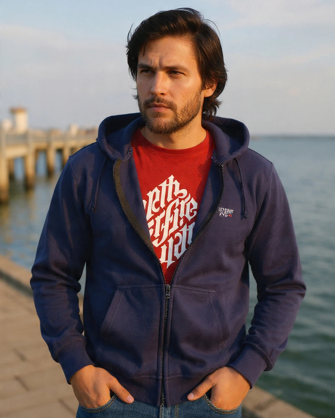 Man wearing a navy blue hoodie with a red shirt underneath, standing by a waterfront.
