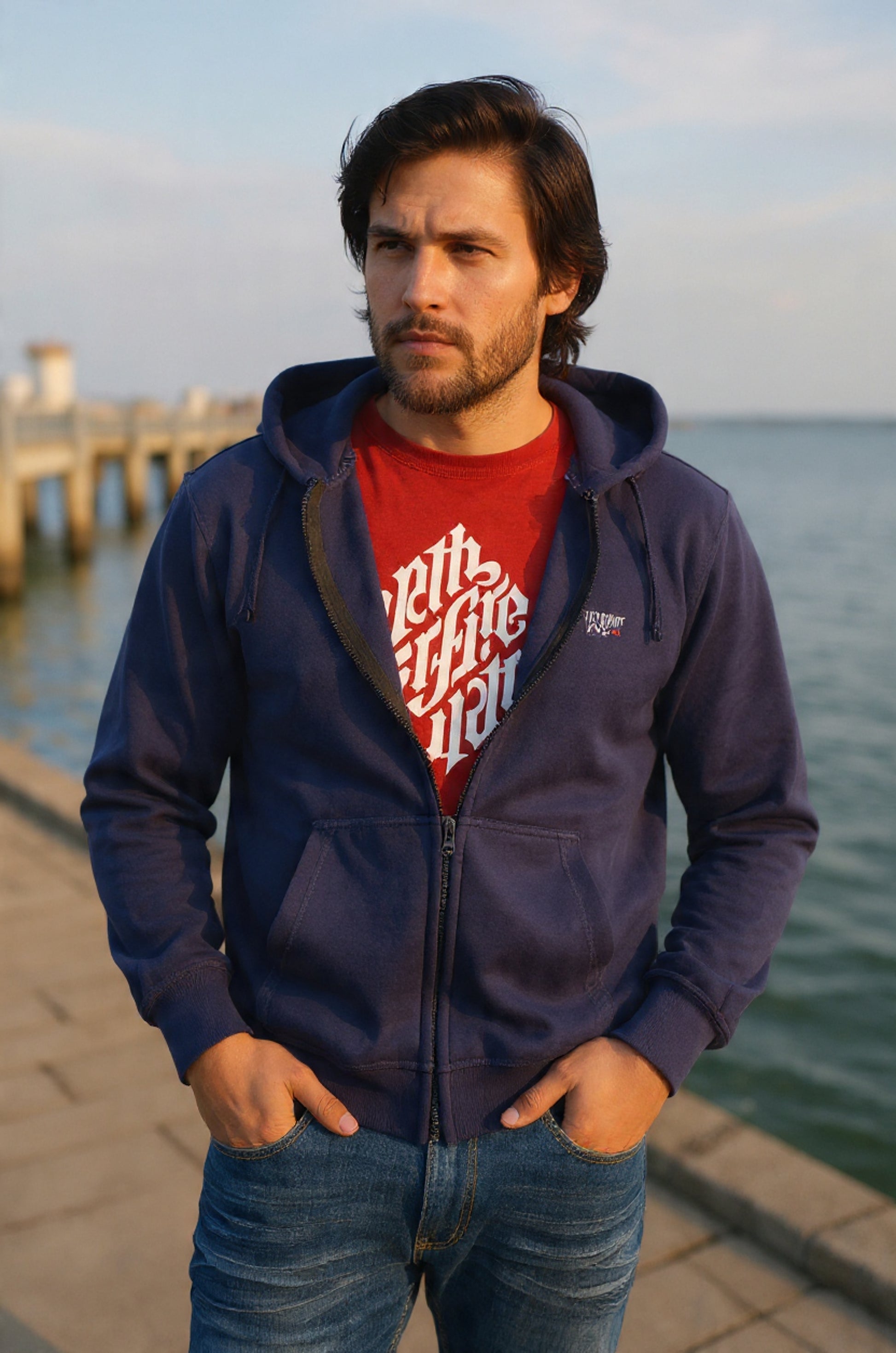 Man wearing a navy blue hoodie with a red shirt underneath, standing by a waterfront.