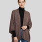 Woman wearing a brown patterned poncho over a black turtleneck and blue jeans on a light gray background