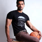 Man sitting on a wooden stool holding a basketball, wearing a black t-shirt with text and brown pants.