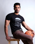 Man sitting on a wooden stool holding a basketball, wearing a black t-shirt with text and brown pants.