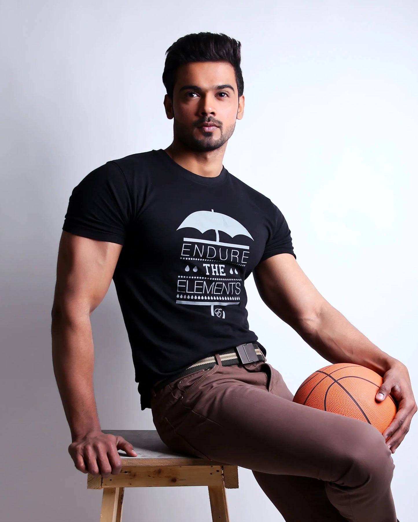 Man sitting on a wooden stool holding a basketball, wearing a black t-shirt with text and brown pants.