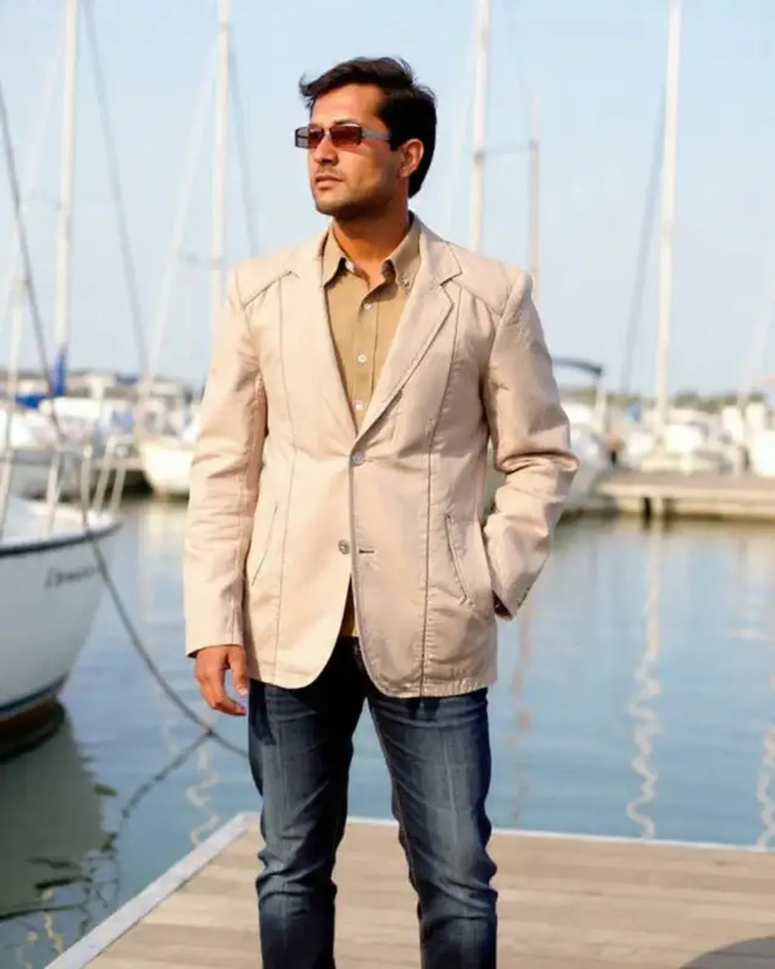 Man in a beige blazer and sunglasses standing on a dock with boats in the background