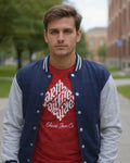 Man wearing a red shirt with white text and a blue and gray varsity jacket on a sidewalk.