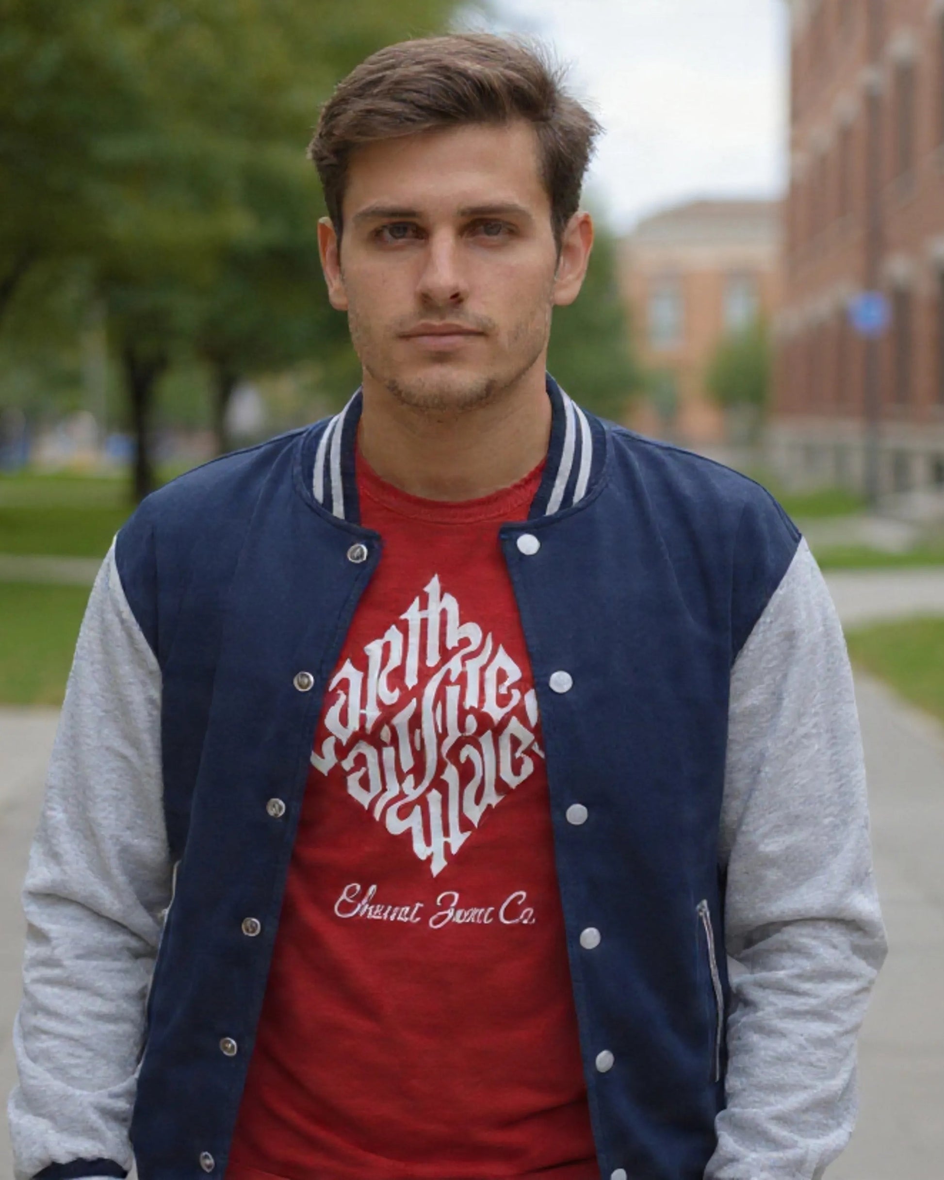 Man wearing a red shirt with white text and a blue and gray varsity jacket on a sidewalk.