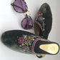 A pair of green velvet mule shoes with floral hand embroidery, accompanied by sunglasses with purple lenses.
