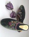 A pair of green velvet mule shoes with floral hand embroidery, accompanied by sunglasses with purple lenses.