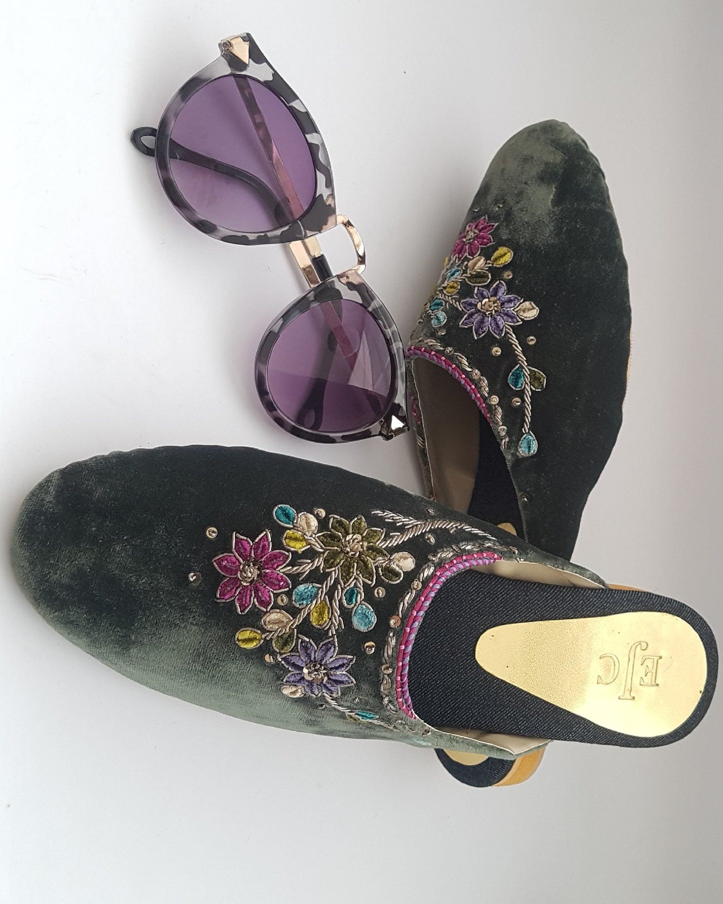 A pair of green velvet mule shoes with floral hand embroidery, accompanied by sunglasses with purple lenses.
