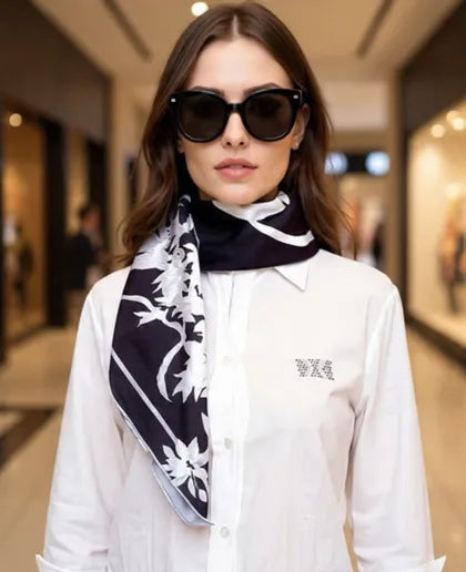 Woman wearing a white shirt and black and white patterned scarf with sunglasses in a blurred indoor setting