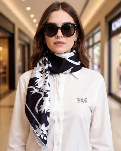 Woman wearing a white shirt and black and white patterned scarf with sunglasses in a blurred indoor setting