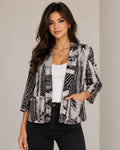 A woman modeling a black and white abstract print blazer style shrug over a white tank top and black pants, standing against a neutral beige background.