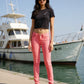Woman wearing a black crop top and pink pants standing by a marina with boats in the background.