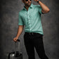 Man in a teal shirt and black pants holding sunglasses and a suitcase against a dark background